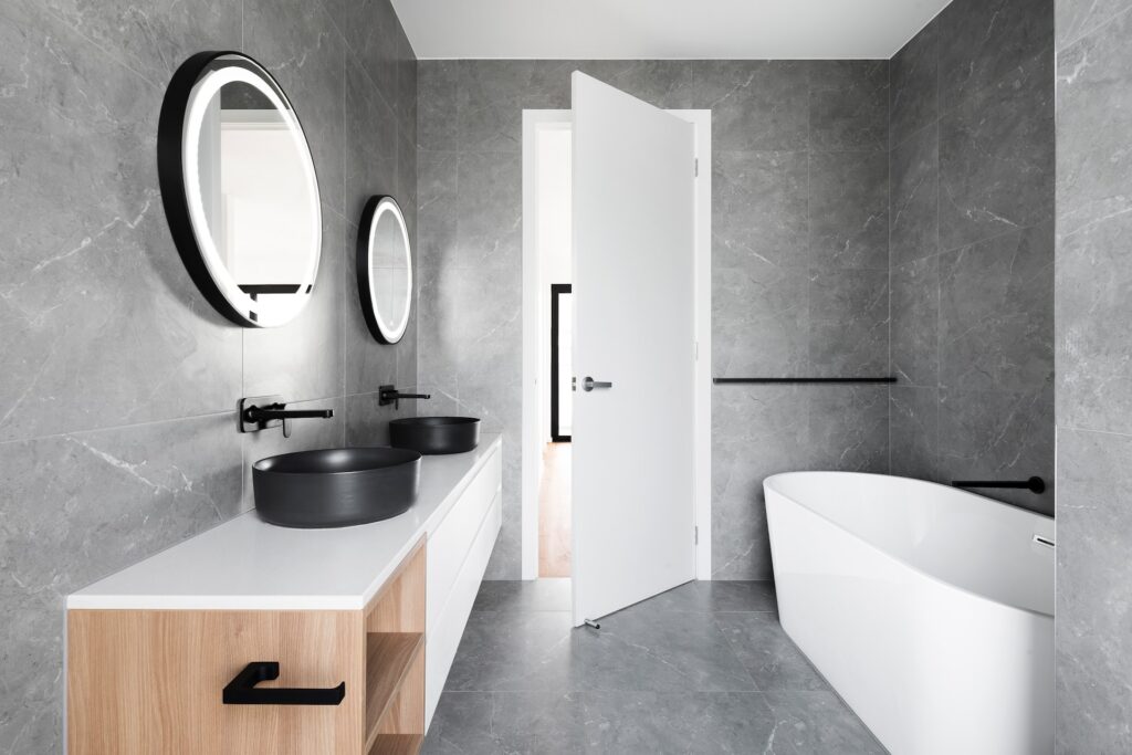 Salle de bain blanche et grise, avec baignoire le long du mur. Accompagné de deux lavabos et de deux miroirs.