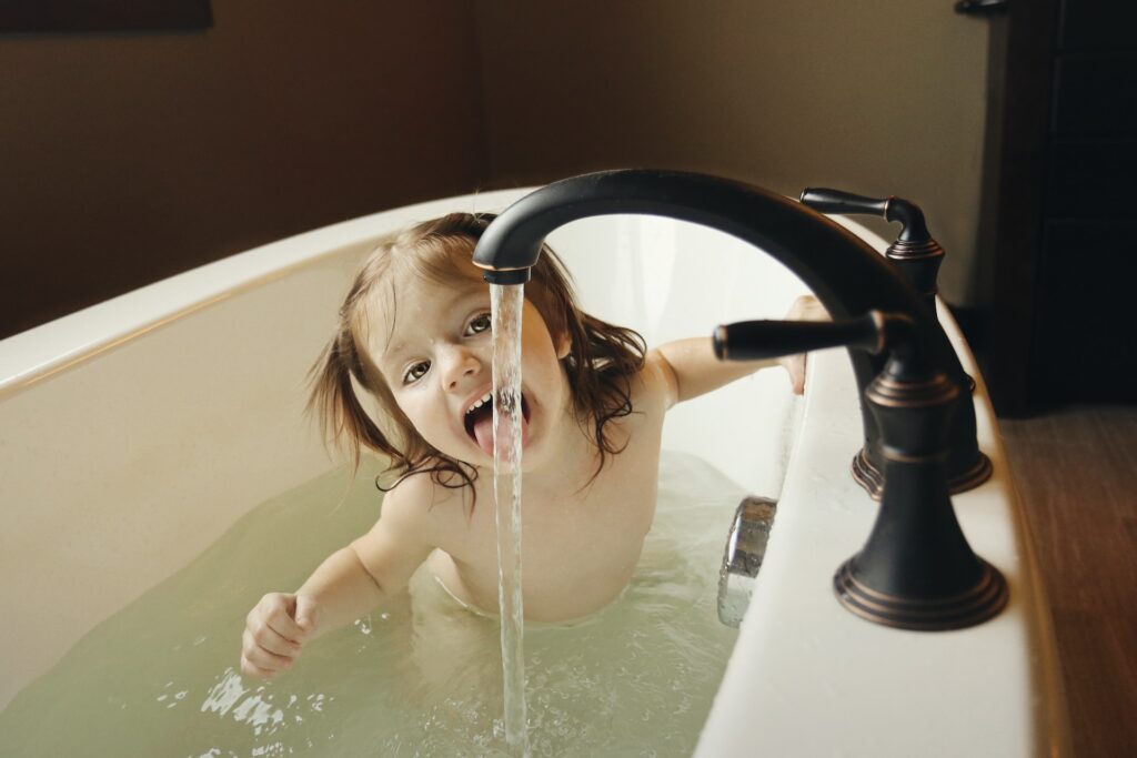 Une petite fille d'un très jeune âge, disons un grand bébé, est dans son bain en train de tirer la langue pour jouer avec le filet d'eau qui coule du robinet noir.
