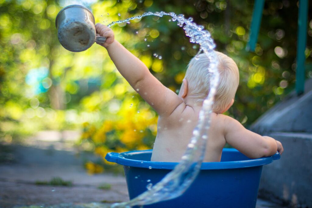 Un bébé est assis dans une bassine bleue située en extérieur. Un joue avec de l'eau qu'il tient dans un pot.