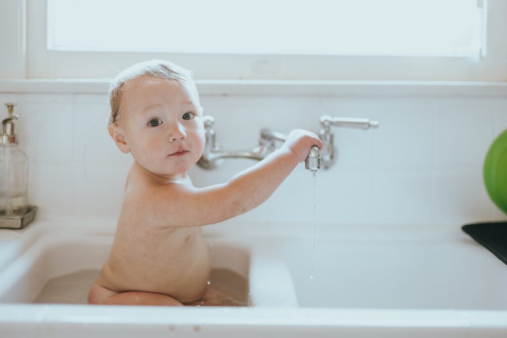 Bébé dans un bain, en train de tenir un robinet des deux mains. L'image est très claire et les murs d'un blanc éclatant.