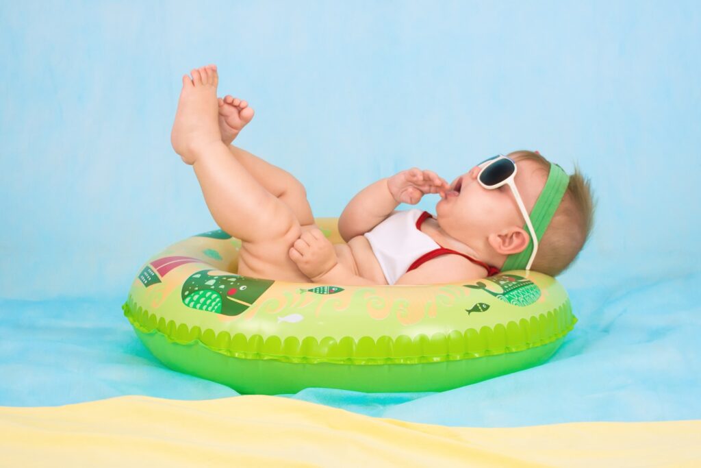 Un bébé est sur une bouée verte dans un décor bleu qui fait penser à une piscine. Il est allongé sur le dos et porte des lunettes de soleil.