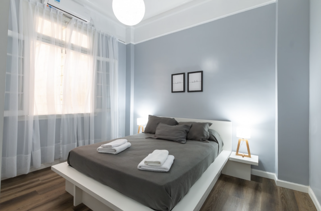 Chambre peinture grise dont la luminosité est voilée par des rideaux blancs. Le lit est en draps sombres comme le sol pour créer un contraste avec les couleurs claires et les lumières de la pièce.