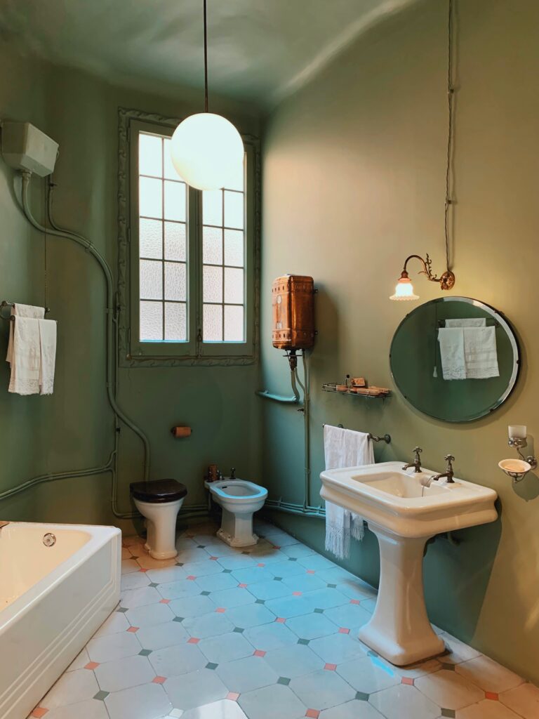 Salle de bain ancienne avec un carrelage en carreaux de ciment blanc et noir. Les murs sont bleu-vert et les meubles blanc et sobre. Le miroir est rond et petit.