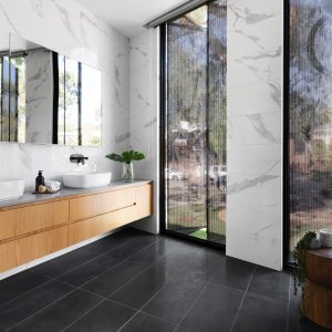 Salle de bain de luxe au mur marbré blanc et au sol en carrelage noir. Le meuble de salle de bain est en bois clair avec deux tiroirs intégrés. Il y'a deux baies vitrées.